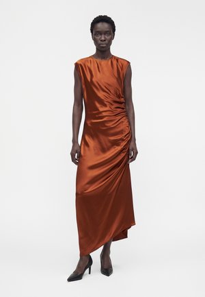 Femme portant une robe sans manches en satin couleur rouille avec fronces sur le côté et ourlet asymétrique, associée à des talons noirs pointus, debout droite.