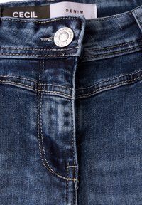 Jeans i denim med en mörkblå tvättning, med en silverfärgad knappstängning och kontrasterande gul sömnad längs sömmarna. Etikett synlig invändigt.