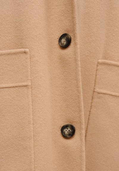 Μπεζ μάλλινο παλτό με απαλή υφή, δύο μπροστινές τσέπες και στρογγυλά σκούρα κουμπιά για κλείσιμο. Κλασικός σχεδιασμός με καθαρές γραμμές.