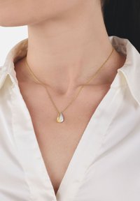Mujer que lleva un collar de cadena dorada con un pequeño colgante en forma de gota bicolor sobre una camisa blanca con cuello.