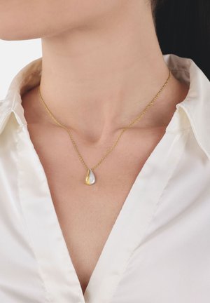 Donna che indossa una collana d'oro con un piccolo ciondolo a forma di goccia bicolore sopra una camicia bianca con colletto.