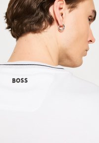 Fehér póló fekete "BOSS" logóval a hátsó részen, lekerekített nyakkal és egy diszkrét fekete díszítéssel a gallérnál.