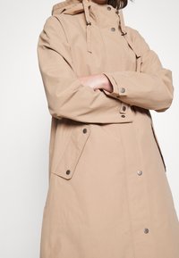 Hellbeiger wasserdichter Mantel mit Kapuze, Druckknöpfen, Seitentaschen und verstellbaren Kordeln. Glatte Textur und lässiges Design.
