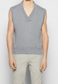 Suéter gris sin mangas con un diseño de cuello en V, que presenta una textura acanalada vertical y un dobladillo con banda. Está hecho de un material de punto suave.