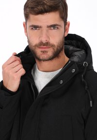 Chaqueta negra con capucha que presenta un cierre de cremallera, botones de presión y un tejido texturizado. La modelo sostiene el cuello, mostrando el forro interior y los detalles del diseño.