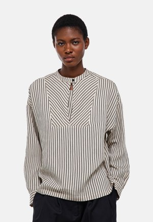 Jeune femme noire portant un chemisier beige à manches longues avec des rayures noires verticales et un petit bouton à l'avant au niveau du cou.