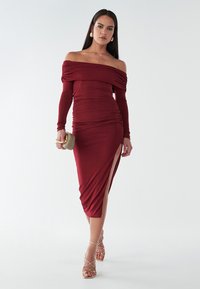 Šaty v burgundskej farbe s odhalenými ramenami, dlhými rukávmi, zúženou textúrou a rozparkom na boku, doplnené o sandále na remienky a malú kabelku.