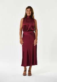 Bordeaux satin kjole med høj hals, indsnørt talje og en flydende nederdel. Sammen med brune remsandaler. Glat tekstur og elegant design.