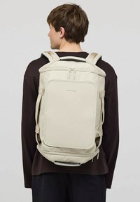 Beige ryggsäck med vadderade axelremmar, rektangulär form, dubbla dragkedjor och framfickdetalj. Slät textur och minimalistisk design.