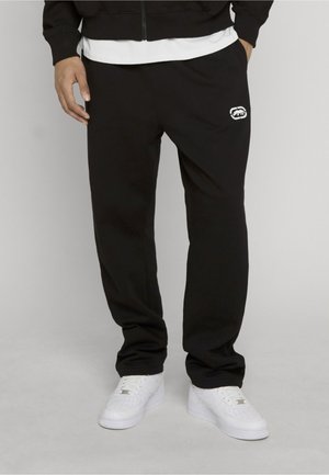 Persona con pantalones de chándal negros con un pequeño logo blanco en el muslo izquierdo y zapatillas blancas, de pie con las manos en los bolsillos.