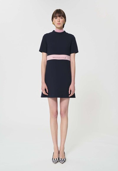 Femme debout portant une robe courte à manches courtes bleu marine avec une ceinture et un col roses, des boucles d'oreilles dorées, et des escarpins pointus à rayures bleu marine et blanches.