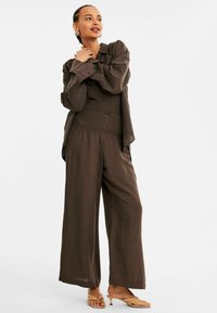 Bruine, lichte, textuurrijke outfit bestaande uit een losse blouse en wijde broek met een smocktail. Neutrale instapschoenen.