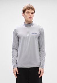 Γκρι μακριά μπλούζα με μακρύ μανίκι και φερμουάρ μέχρι το ¼. Διαθέτει υφή υφάσματος και ένα μπλε λογότυπο Calvin Klein στο στήθος. Λείο φινίρισμα.