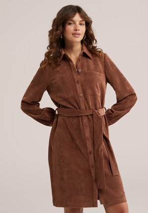 Femme aux cheveux bouclés portant une robe en velours côtelé marron boutonnée avec ceinture, debout les mains sur les hanches devant un fond uni.