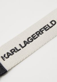 Etichetta in tela di colore crema con scritta in rilievo nera che dice "KARL LAGERFELD." I bordi sono rifiniti con un accento in pelle nera.