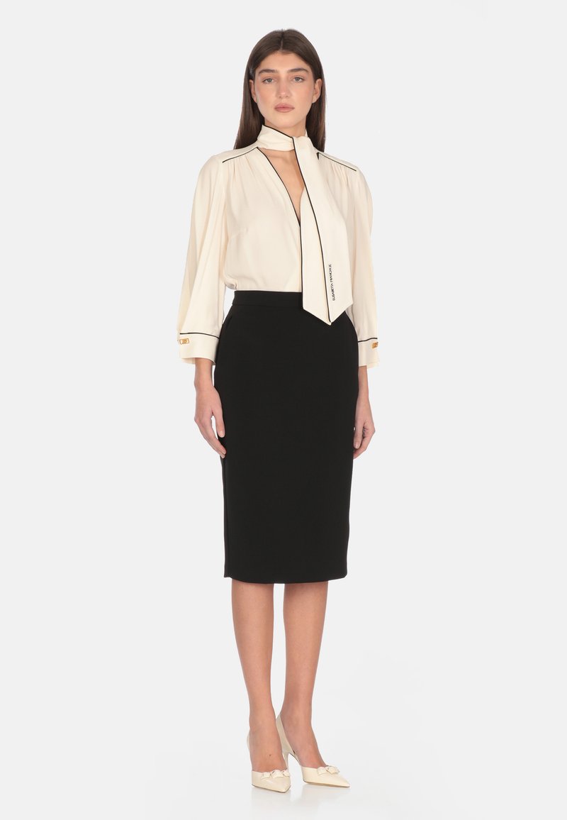 Blusa avorio con bordi neri, maniche lunghe e una sciarpa annodata; abbinata a una gonna a tubino nera aderente. Tacchi crema con dettaglio a fiocco.