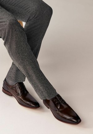 Chaussures habillées en cuir marron avec un bout pointu, un laçage et des coutures décoratives, assorties à un pantalon texturé gris foncé.