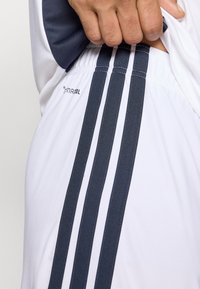 Λευκοί αθλητικοί σορτς με τρεις κάθετες σκούρες μπλε ρίγες, φτιαγμένοι από μαλακό ύφασμα, με ένα λογότυπο στο πλάι.