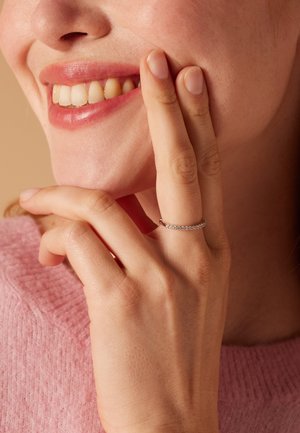 Ring featuring a delicate band set with small clear stones, worn on a woman's hand touching her face, with a pink knit sweater in the background.