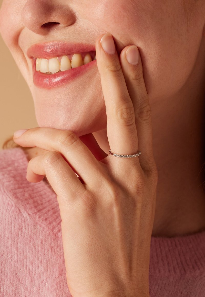 Ring mit einem zarten Band, das mit kleinen klaren Steinen besetzt ist. Er wird an der Hand einer Frau getragen, die ihr Gesicht berührt, mit einem rosa Strickpullover im Hintergrund.