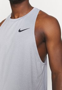Camiseta deportiva sin mangas de color gris claro, con un escote redondo y grandes sisas. Presenta el logotipo de Nike en negro en el pecho.