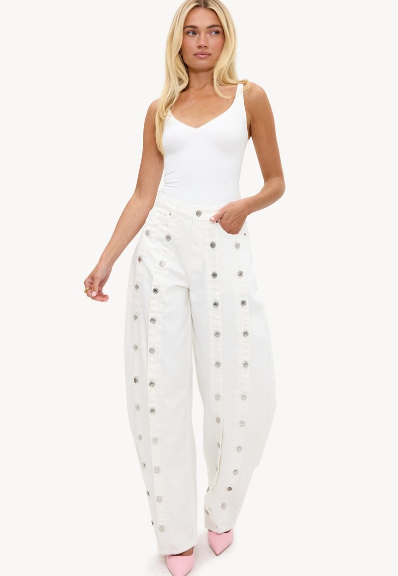 Femme blonde portant un haut blanc sans manches, un pantalon blanc taille haute à jambes larges avec des boutons métalliques, et des talons roses pointus.