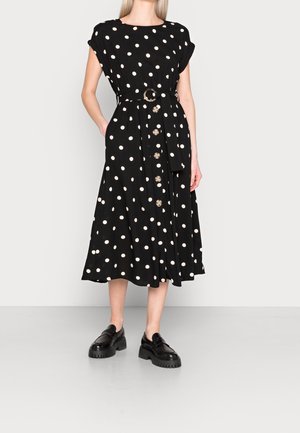 Nainen, jolla on päällään musta midi-mekko, jossa valkoiset pallokuvat, napit edessä, vyötäröllä vyö sekä mustat paksupohjaiset loafersit, seisoo yksivärisen taustan edessä.
