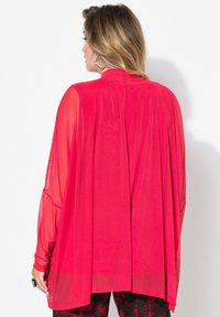 Rote, leichte, durchsichtige Kimono-Bluse mit langen Ärmeln, offenem Rücken und strukturierten Details, kombiniert mit gemusterten Hosen.