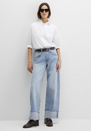 Weißes Hemd mit Knöpfen, hellblaue Jeans mit weitem Bein und gekürzten Säumen, schwarzer Gürtel und übergroße schwarze Sonnenbrille. Schwarze Schuhe vervollständigen das Outfit.