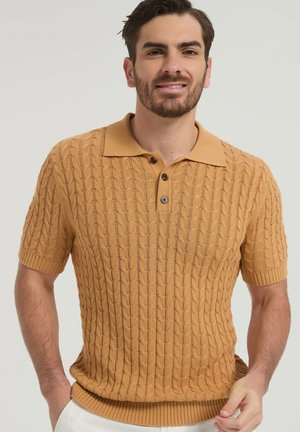 Homme barbu portant un polo jaune moutarde en maille torsadée à manches courtes, debout avec une main dans la poche, sur un fond uni.