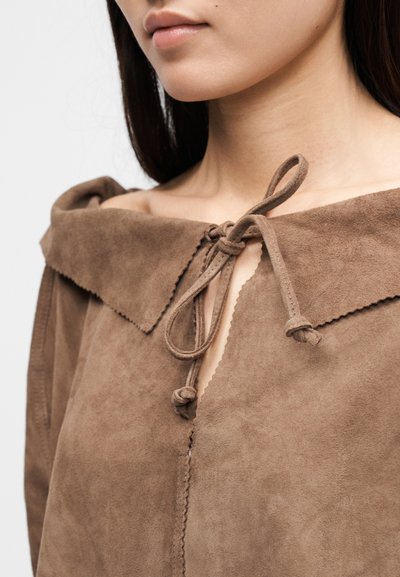 Καφέ αμάνικο από σουέντ με φαρδιά γιακά, διαθέτει δέσιμο στο λαιμό και κυματιστές άκρες κατά μήκος του γιακά και της πατιλέτας.