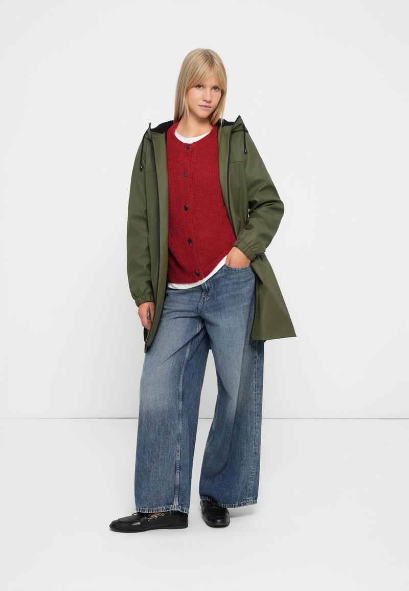 Groene waterdichte jas met een capuchon, rode gestructureerde trui, brede blauwe spijkerbroek en zwarte schoenen. Eenvoudig ontwerp en casual pasvorm.