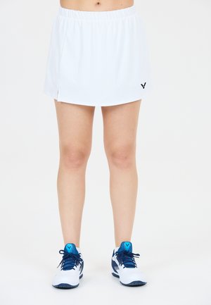 Kvinde iført hvid sportsskjørt og hvide sneakers med blå detaljer, stående mod en ensfarvet hvid baggrund.