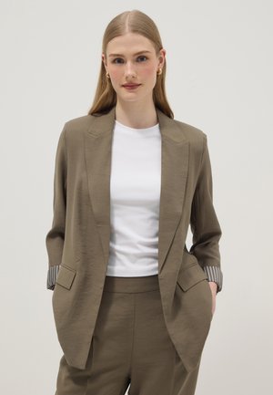 Vrouw met lang blond haar draagt gouden oorringen, een olijfgroen pak met opgerolde mouwen en een wit shirt met ronde hals, handen in de zakken.