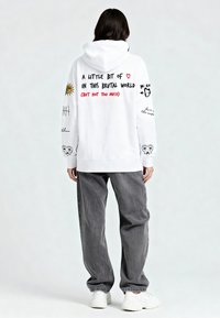 Felpa con cappuccio bianca con varie stampe grafiche, tra cui testi e illustrazioni, sulla schiena e sulle maniche. Abbinata a pantaloni grigi e sneakers bianche.