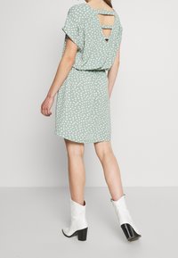 Femme portant une robe vert menthe à pois blancs, avec un dos nu, et des bottes à talons blanches mi-mollet, debout sur un sol gris.