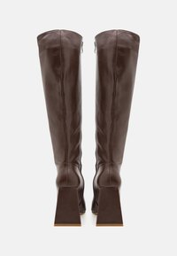 Bottes hautes en cuir marron avec talons carrés, vues de dos sur un fond blanc uni.
