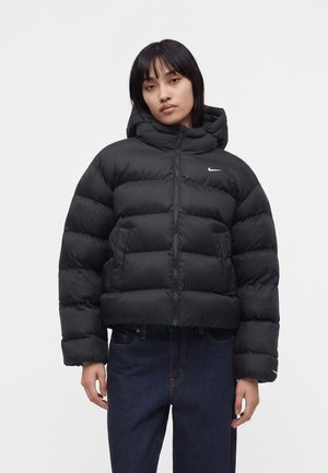 Ung kvinde iført en sort dunjakke med hætte og lille hvid Nike-logo, kombineret med mørkeblå jeans, stående mod en neutral baggrund.
