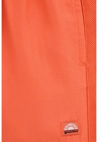 Oranžne športne kratke hlače z gladkim materialom in mrežastim stranskim panelom. Imajo všito logotipno nalepko z napisom »Chasing Waves«.