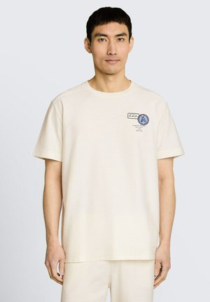 Man draagt een crèmekleurig T-shirt met korte mouwen en een klein blauw-zwart logo op de linkerborst, staand tegen een effen achtergrond.