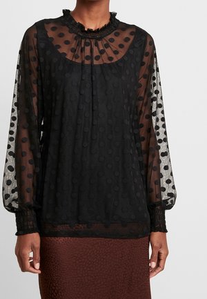 Czarna prześwitująca bluzka z długim rękawem z podniesionymi grochami, marszczonym dekoltem i elastycznymi mankietami, noszona z brązową teksturowaną spódnicą.