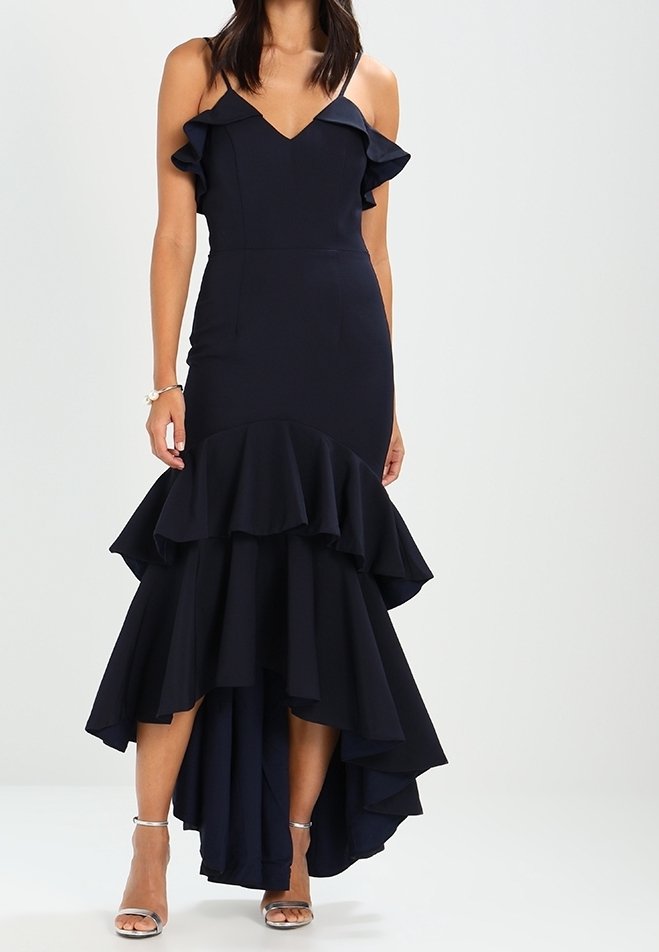Femme portant une robe sans manches bleu marine avec des volants en couches et un ourlet asymétrique haut-bas, associée à des talons ouverts argentés.