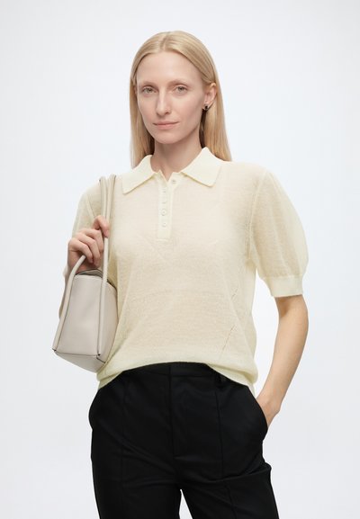 Femme blonde portant un polo en maille crème à manches courtes et un pantalon noir, tenant un sac à main beige clair sur son épaule.