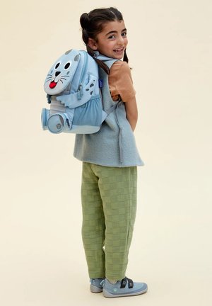 Smiling child with braided hair wearing a blue animal-themed backpack, orange shirt, gray vest, green pants, and gray shoes standing sideways.