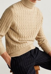 Homme portant un pull à col roulé beige tricoté en torsades et un pantalon foncé à rayures fines, assis sur un tabouret en bois les mains dans les poches.