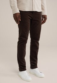 Smeđi kordura hlače slim fit kroja, s glatkom teksturom. Uparene s bijelim tenisicama, naglašavajući čist, ležeran izgled.