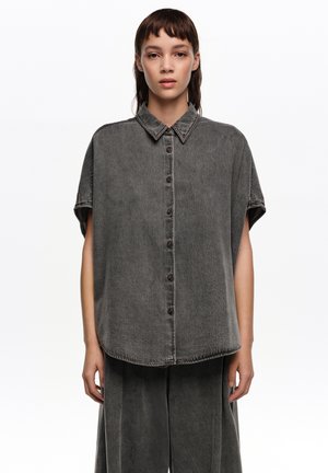 Mujer con camisa negra lavada de manga corta y botones, suelta, y pantalones anchos a juego, de pie contra un fondo blanco.