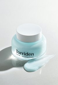 Torriden Soothing Cream läpinäkyvässä lasipurkissa, jossa on valkoinen kansi. Vaaleansininen voide pursuaa purkista, paljastaen sileän ja kiiltävän koostumuksen.