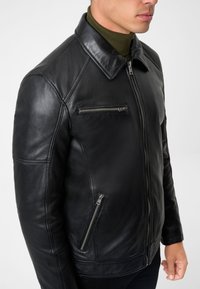Mann trägt schwarze Lederjacke mit vorderen und Brust-Reißverschlusstaschen über einem olivgrünen Rollkragenpullover.