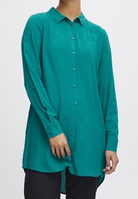 Chemise boutonnée couleur sarcelle, manches longues, col, tissu doux, ourlet arrière légèrement plus long, avec de petits boutons et une texture lisse.